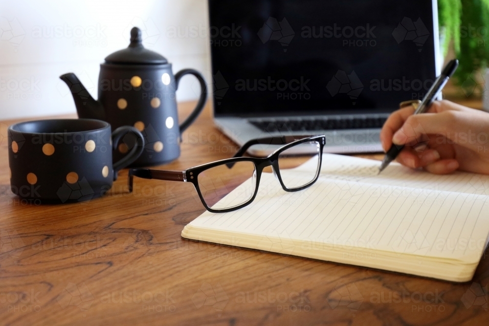 Detail shot of a hand writing in a notebook with laptop and teapot in background - Australian Stock Image