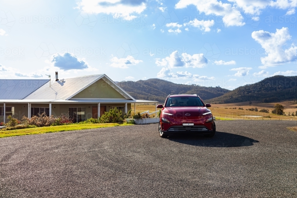 Driving red electric vehicle past country farm house on hilltop - Australian Stock Image