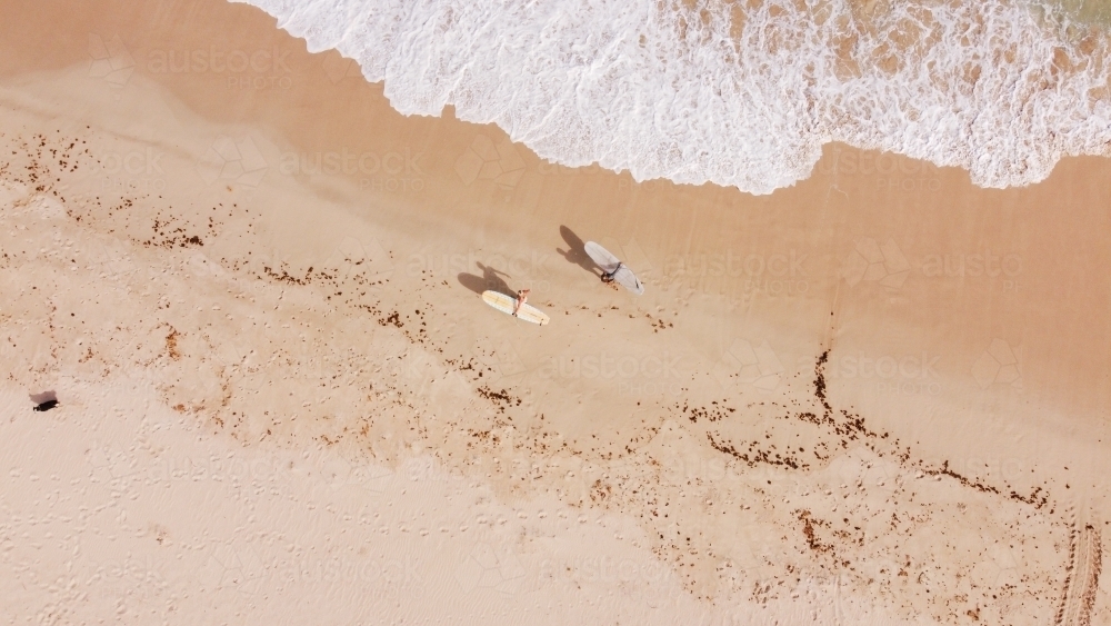 Drone shot of two surfers carrying longboards and walking on sandy beach - Australian Stock Image