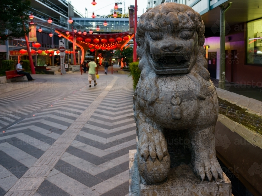 Early evening photo of lion statue in Chinatown with patterned paving and red lanterns - Australian Stock Image