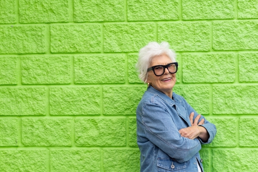 elderly lady in denim jacket standing against a lime green wall - Australian Stock Image