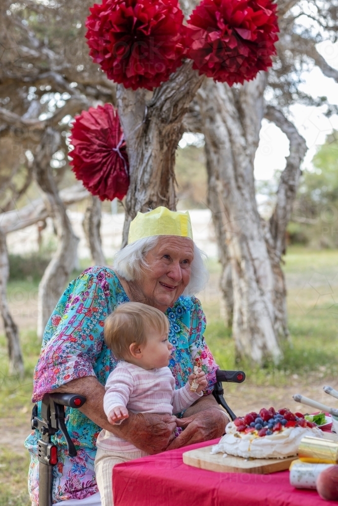 elderly lady wearing paper crown and sitting in walker chair with baby on her lap - Australian Stock Image