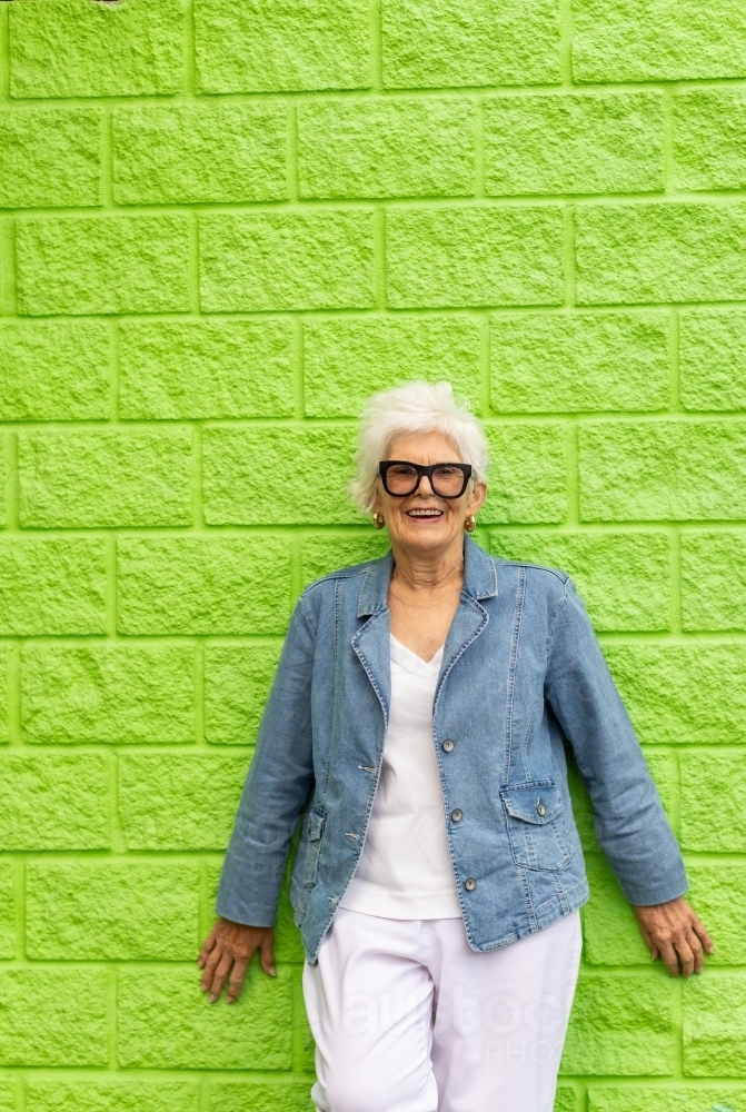 elderly woman in white clothes and denim jacket standing against a bright lime green wall - Australian Stock Image