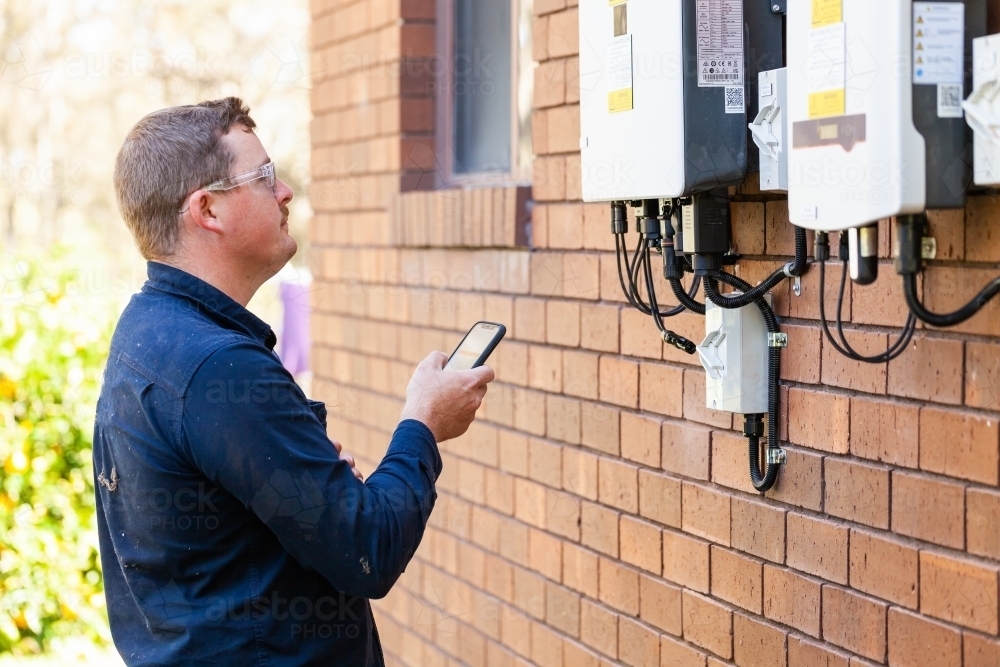 Electrician troubleshooting solar power box on wall of home using phone - Australian Stock Image