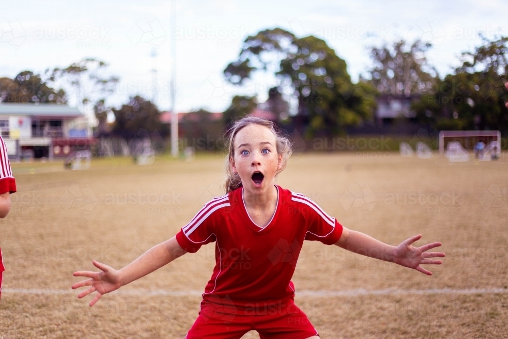 Excited tween girl in a red football uniform yelling with her arms outstretched - Australian Stock Image