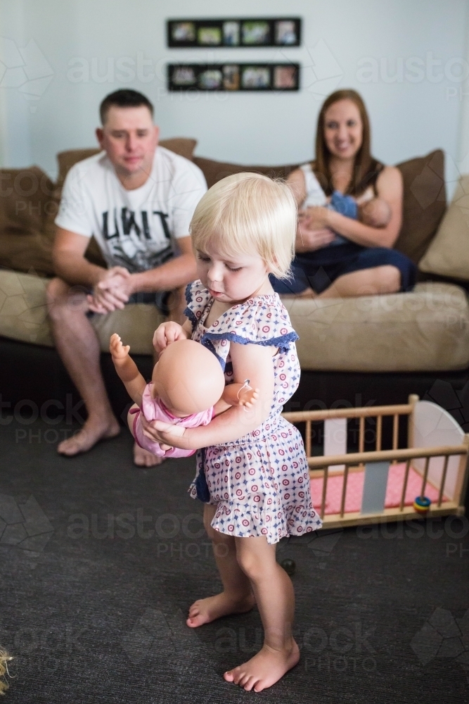 Family at home sitting on lounge with mother feeding newborn baby watching daughter play with doll - Australian Stock Image