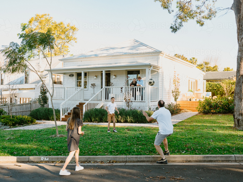 Family playing soccer in front yard of white weatherboard home in suburbs - Australian Stock Image