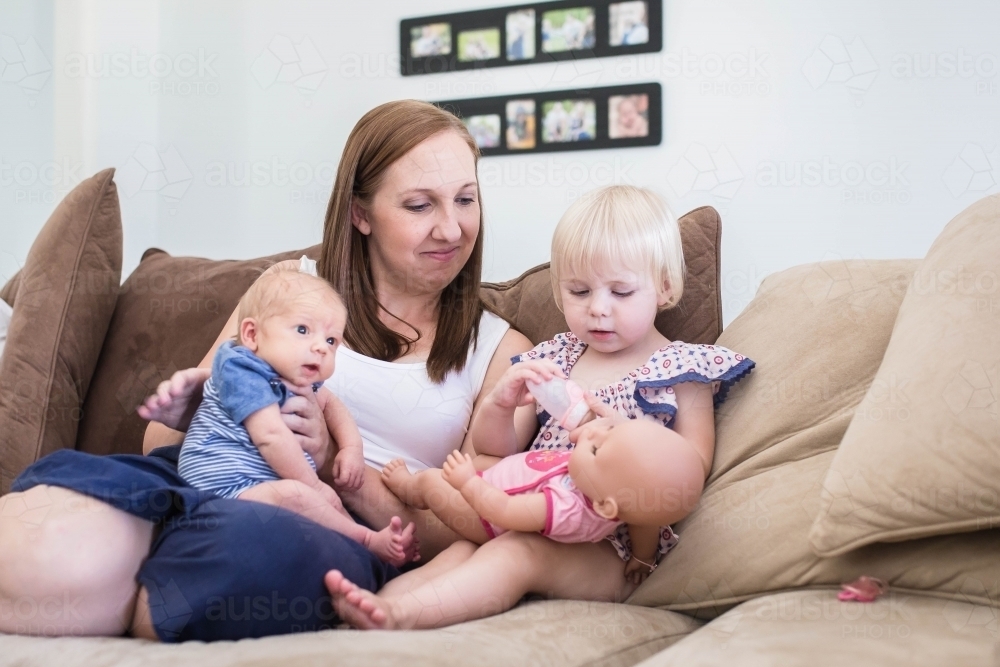 Family sitting on lounge with mum burpring newborn son while daughter plays with doll - Australian Stock Image