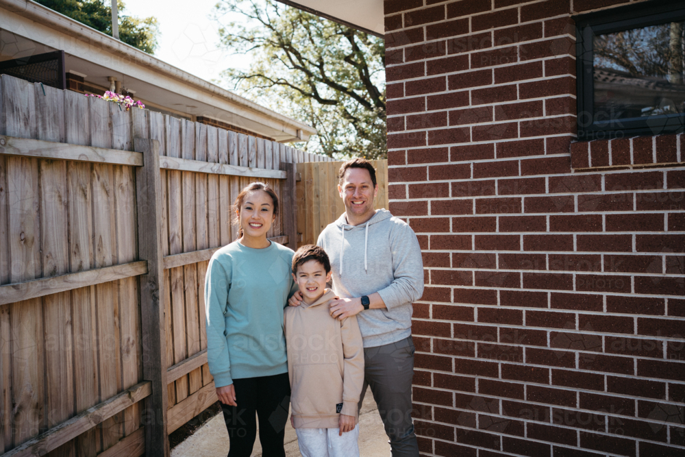 Family standing in front of the brick wall of their house. - Australian Stock Image