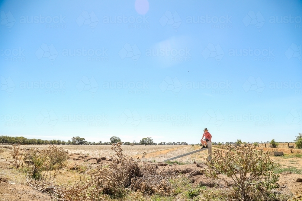 Farm boy sitting on fence of rural property in dry summer drought conditions - Australian Stock Image