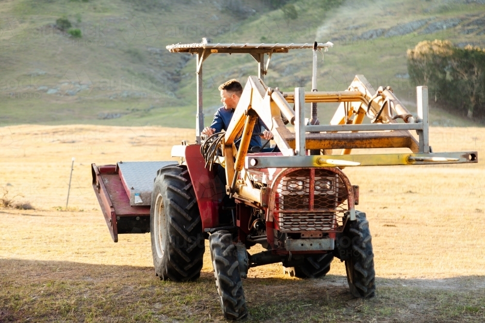 Farmer backing tractor with slasher and forklift attachments - Australian Stock Image