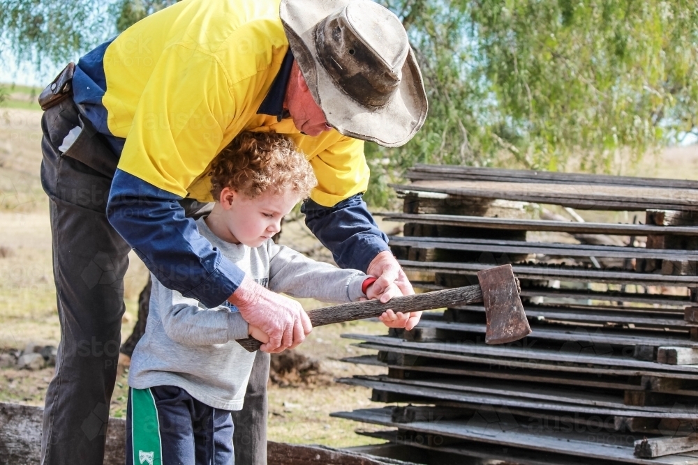 Farmer showing child how to hold axe to chop wood - Australian Stock Image