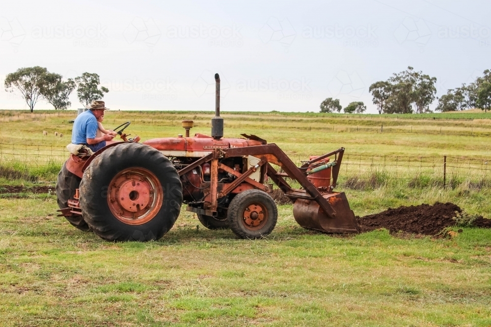 Farmer with grandchild on lap sitting on tractor in farm paddock scooping dirt - Australian Stock Image