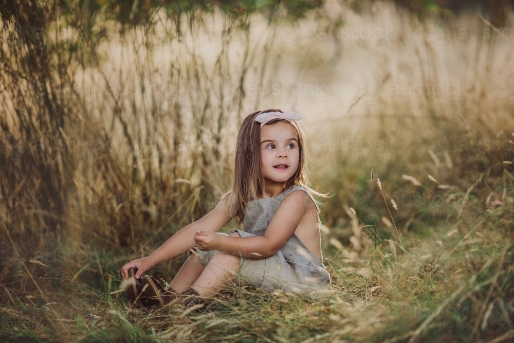 Fashionable young girl, sitting amongst long grass in a farm setting - Australian Stock Image