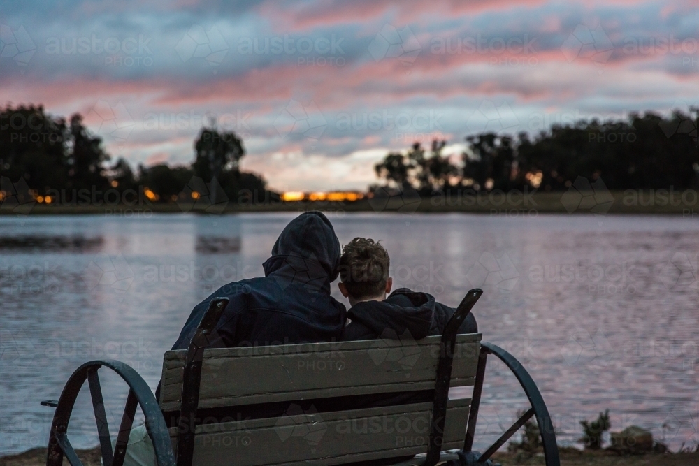 Father and son sitting on chair looking out over river water at sunset - Australian Stock Image