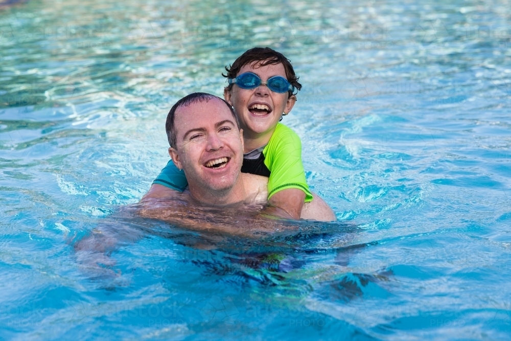 Father and son swimming playing in pool with boy's arms around neck laughing - Australian Stock Image