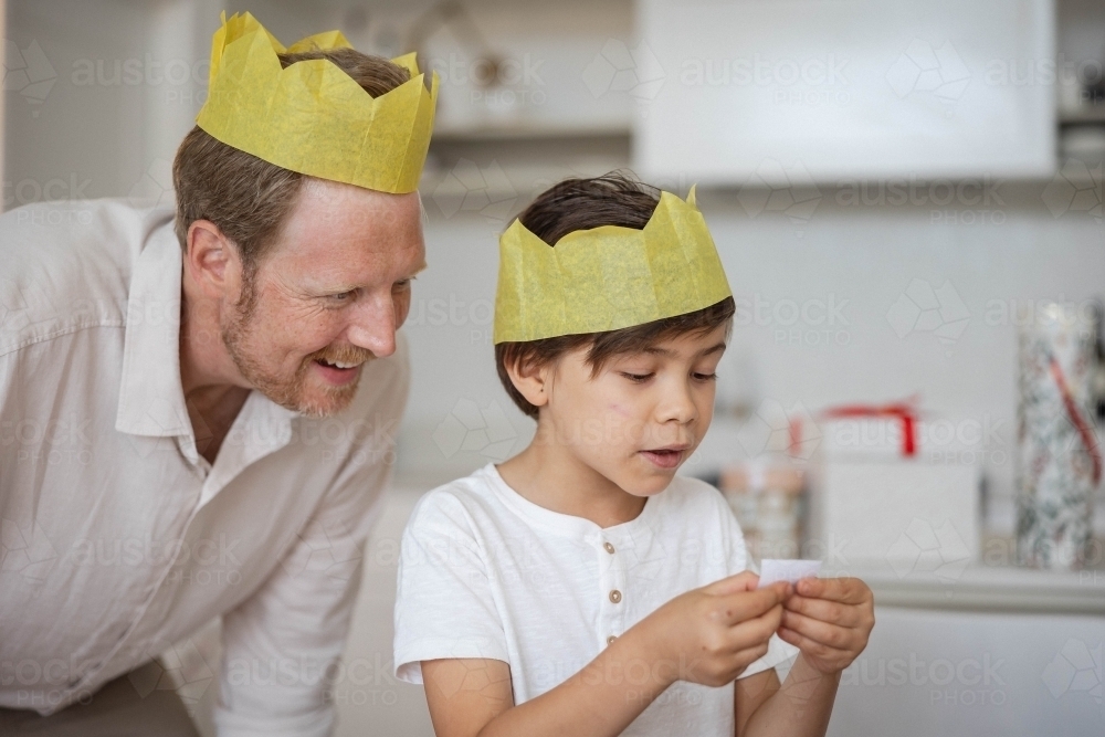 Father and son wearing yellow crowns reading Christmas cracker joke - Australian Stock Image