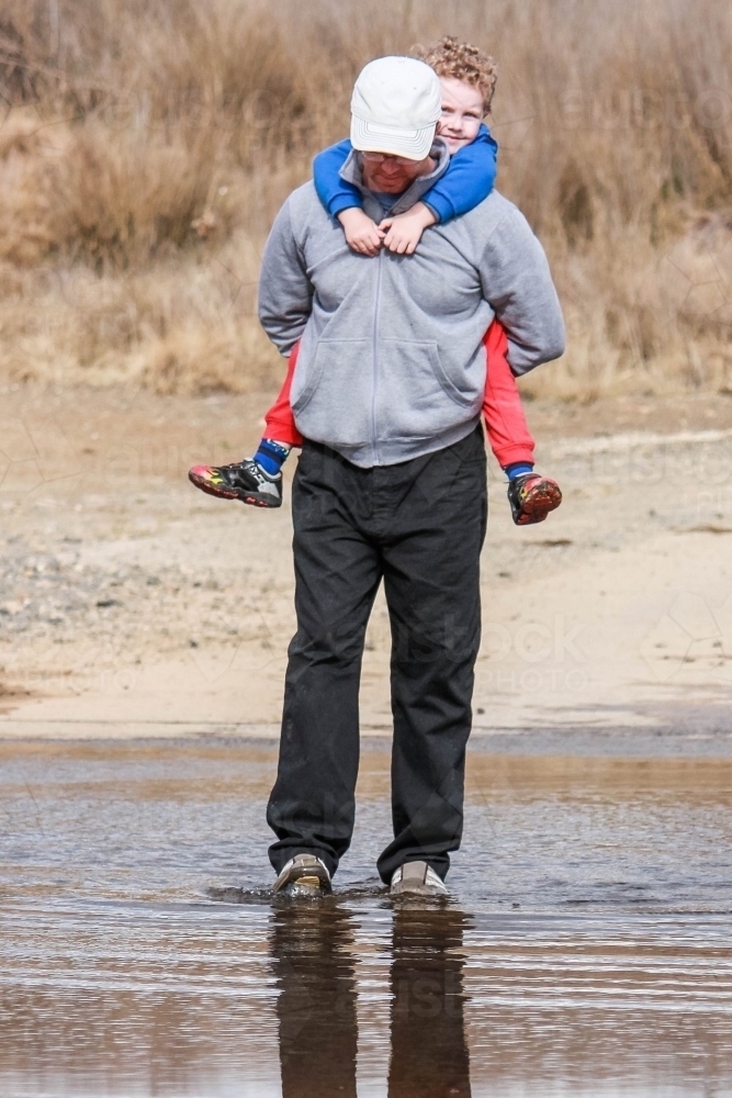 Father carrying son piggy back across water on road - Australian Stock Image