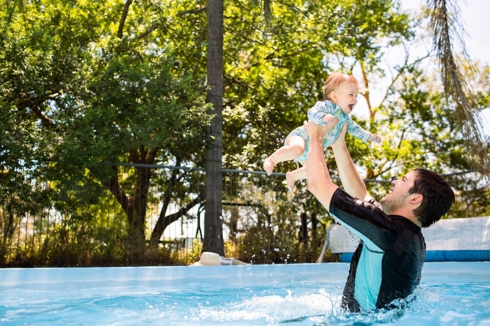 Father playing with baby in swimming pool dad lifting child high into air splashing in water - Australian Stock Image