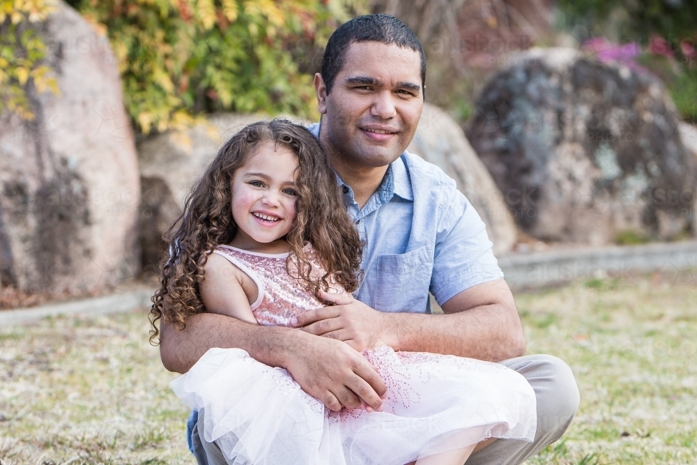 Father sitting with mixed race aboriginal caucasian daughter on lap in garden - Australian Stock Image