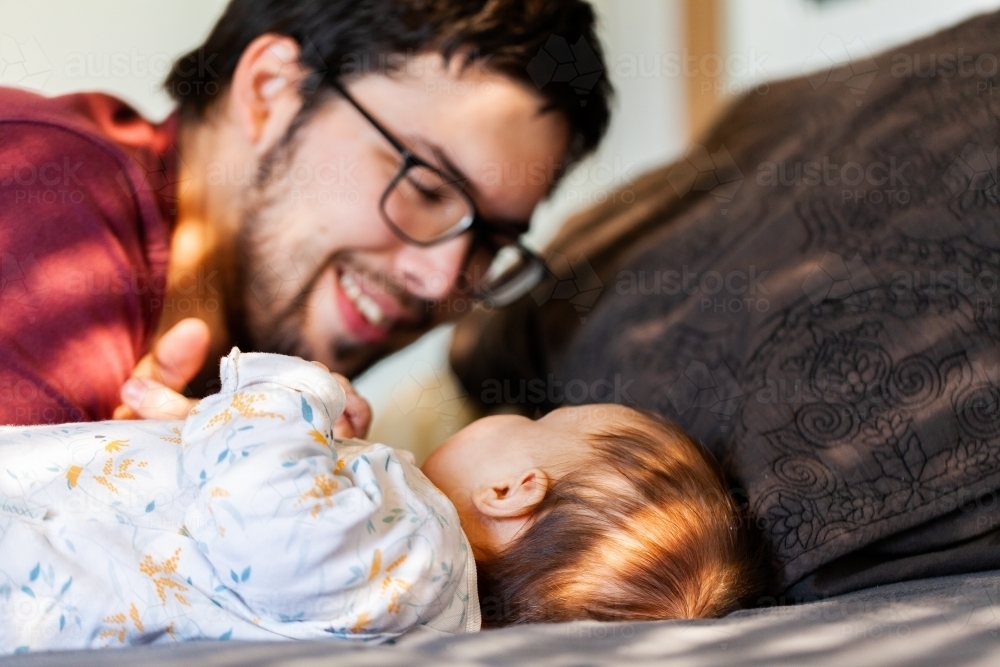 Father smiling and interacting with baby in morning sunlight on bed - Australian Stock Image