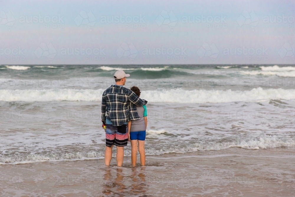 Father standing with arm around son looking out at waves on beach at sunset - Australian Stock Image