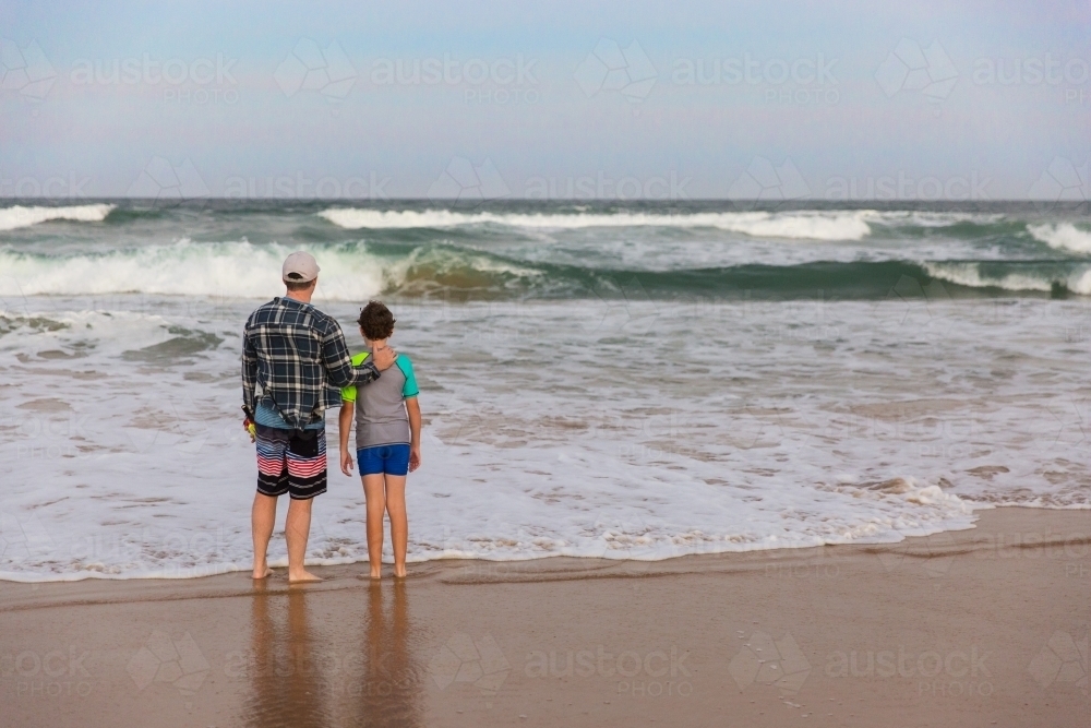 Father standing with hand on son's back at beach looking at waves - Australian Stock Image