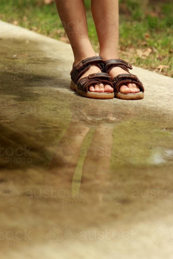 Feet and shoes of a young boy reflected in a puddle on the footpath - Australian Stock Image