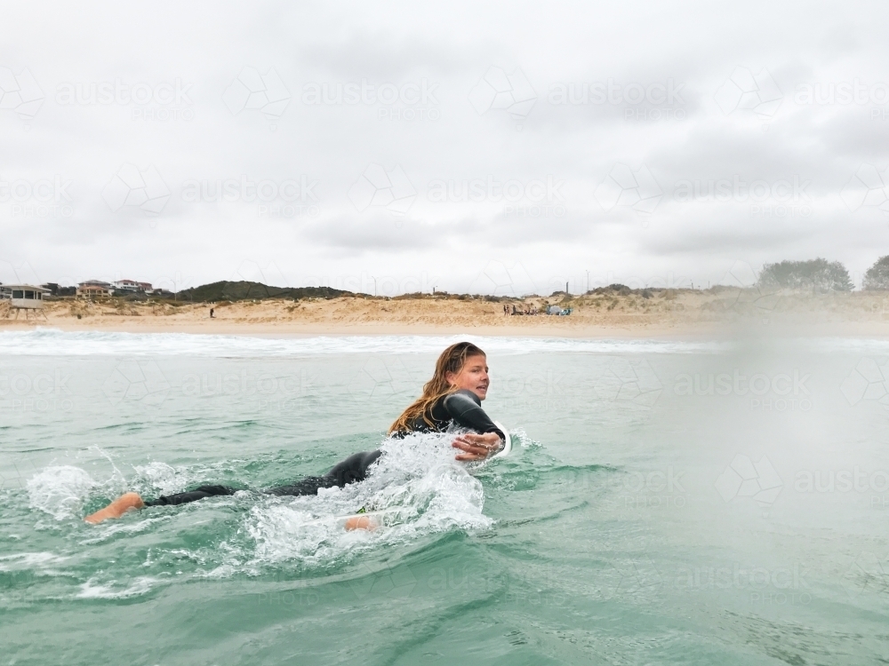 female paddling on surfboard looking back into waves with beach in background on overcast day - Australian Stock Image