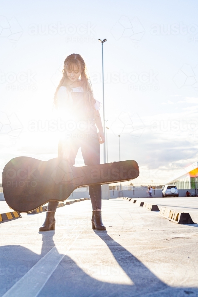 Female teenager posing in empty carpark with guitar case - Australian Stock Image