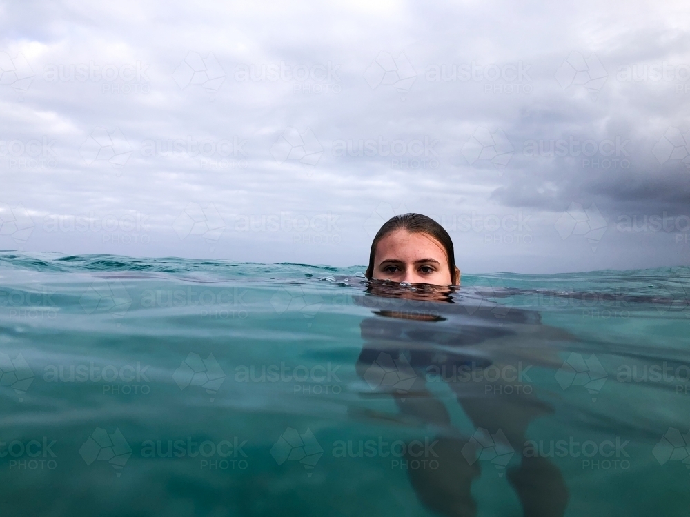 Female teenager with head semi submerged floating in ocean on glassy overcast day - Australian Stock Image