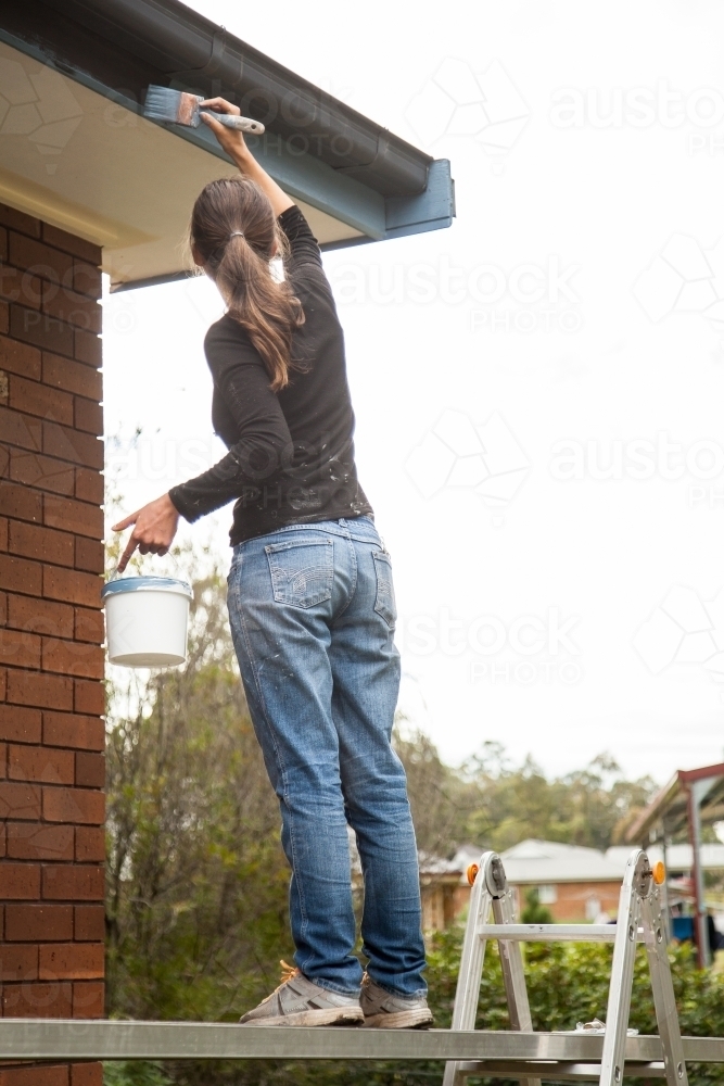 Female tradeswoman painting bargeboards of home on overcast day - Australian Stock Image