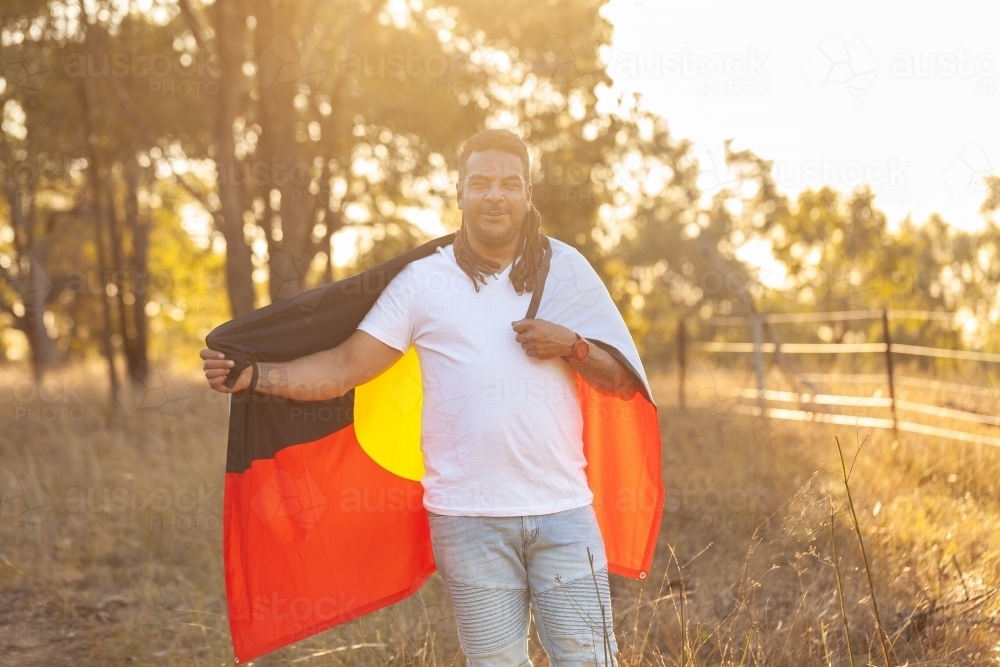 First Nation Australian man standing with Aboriginal flag in bushland - Australian Stock Image