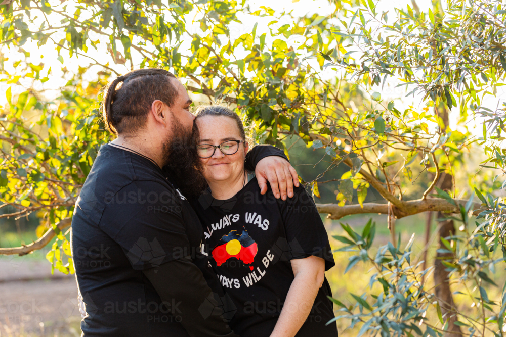 First Nations Australian couple embrace and husband kissing wife in Aussie bushland at sunset - Australian Stock Image