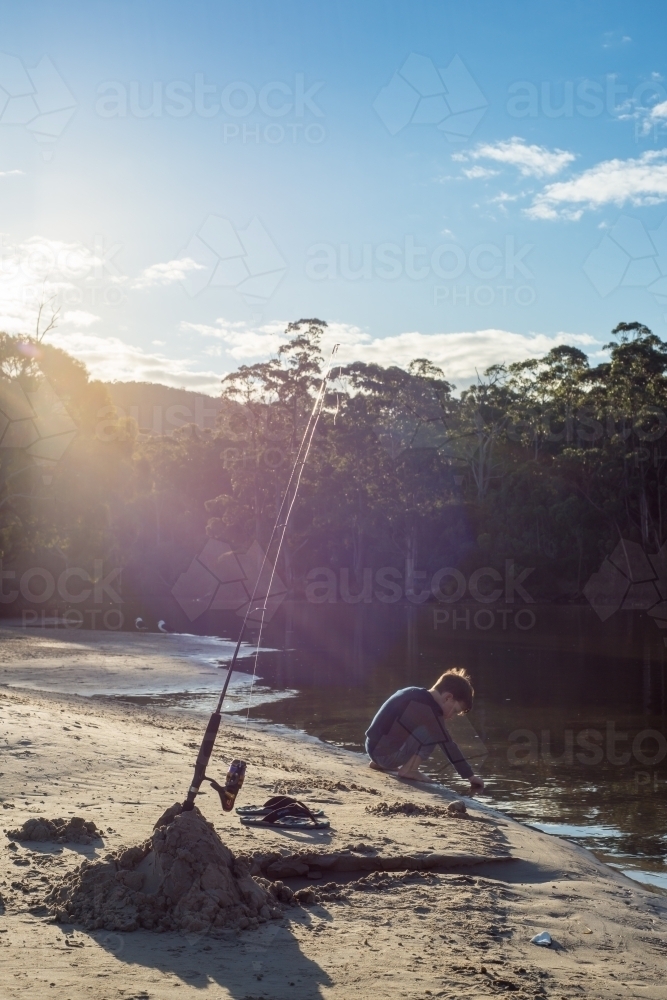 Fishing rod and boy playing in river outlet with sun flare trees and blue sky - Australian Stock Image