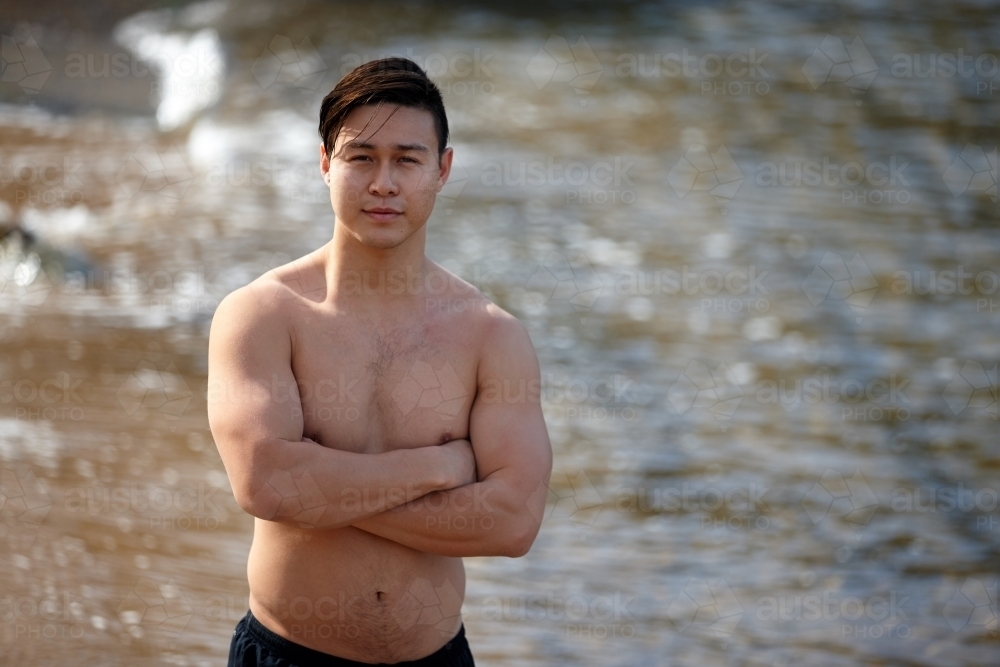 Fit Asian man folding arms at beach wearing only shorts - Australian Stock Image