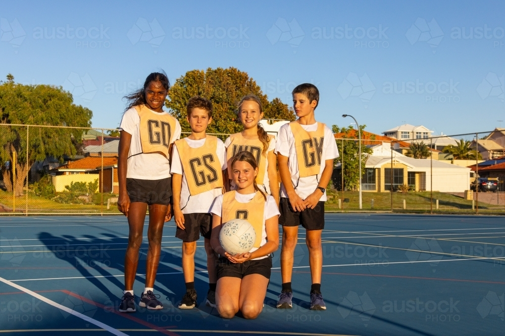 five children from netball team gathered together on blue playing surface for photo - Australian Stock Image