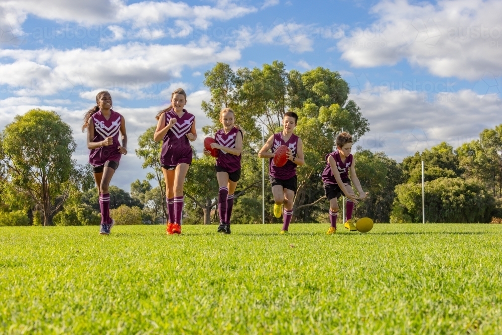 five children in football uniforms running towards the camera on green grass - Australian Stock Image