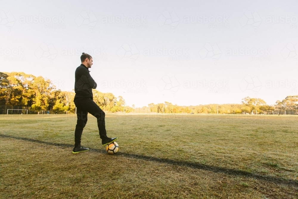 Full body shot of a young man standing on a big field stepping on a soccer ball with one foot - Australian Stock Image