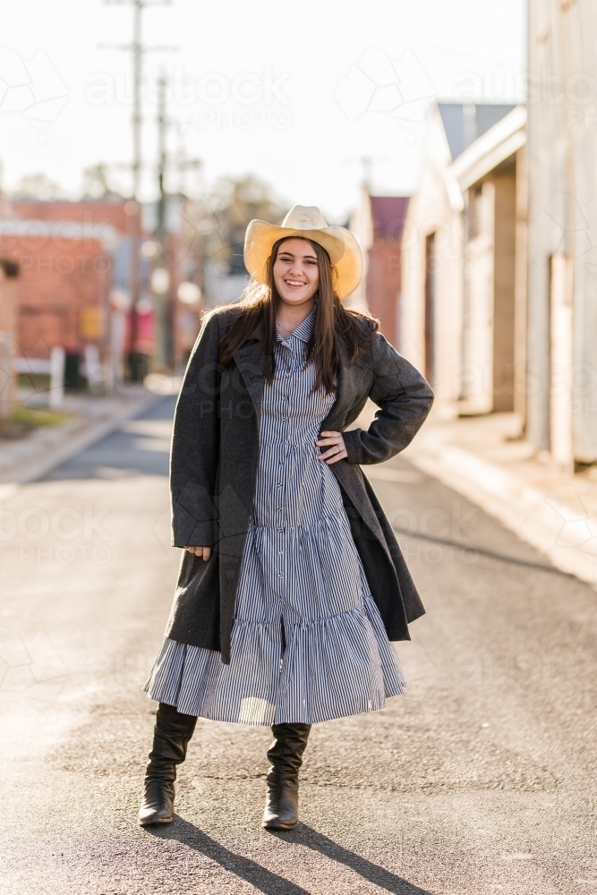 Full length young woman standing with hand on hip wearing boots and hat smiling - Australian Stock Image