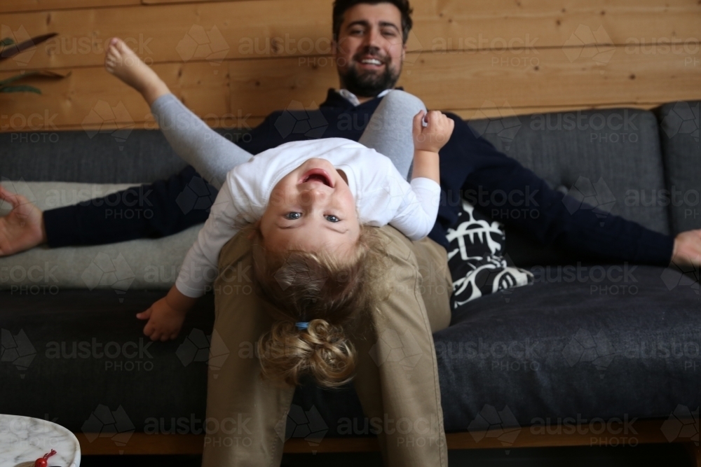 Girl lying on dad's lap on lounge looking at camera - Australian Stock Image