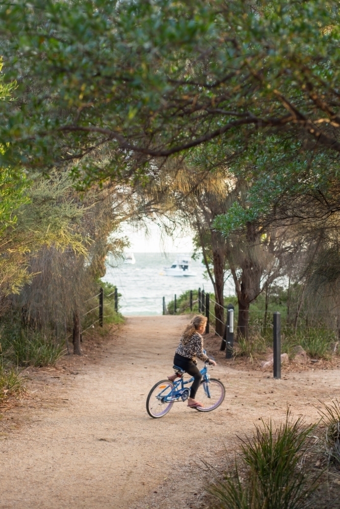 Girl riding a bike on gravel surrounded by trees and beach - Australian Stock Image