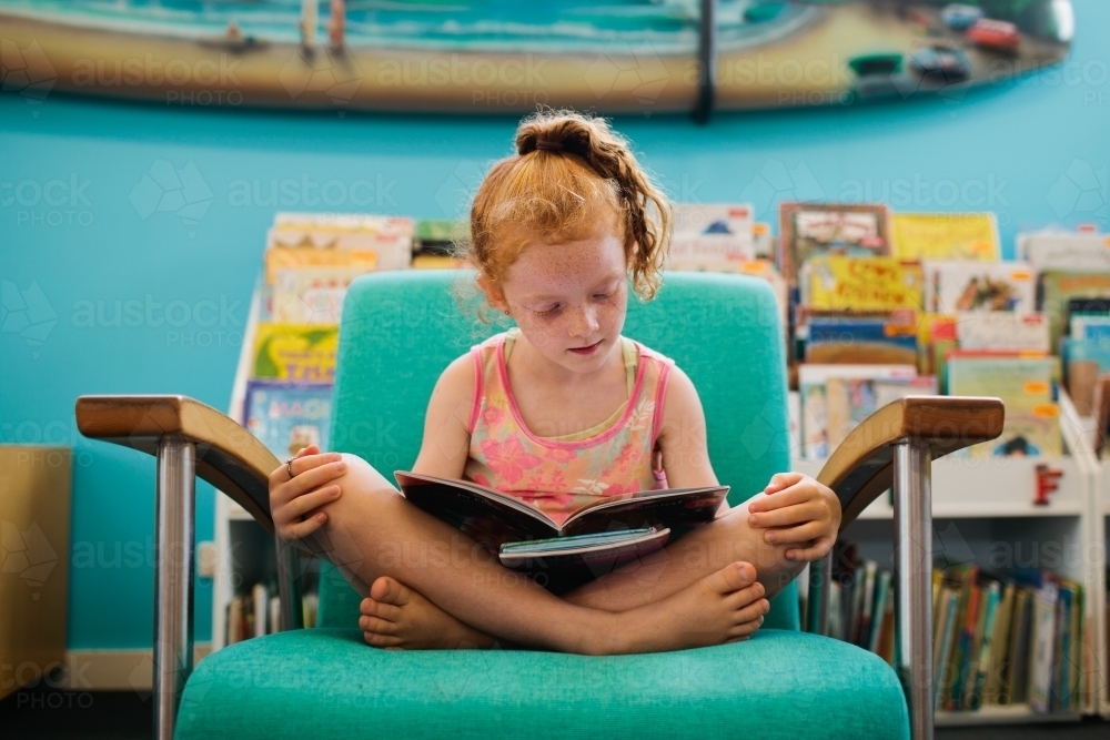 Girl sitting on a chair reading a book in a Library - Australian Stock Image