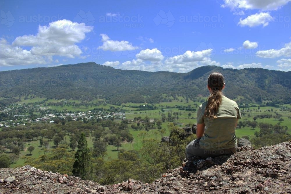 Girl sitting on a rocky ledge overlooking hills and valley - Australian Stock Image