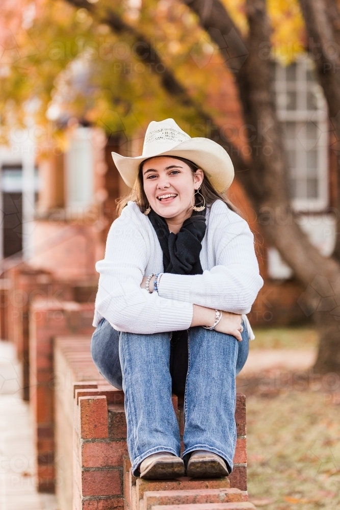 Girl sitting on brick wall smiling hugging knees - Australian Stock Image