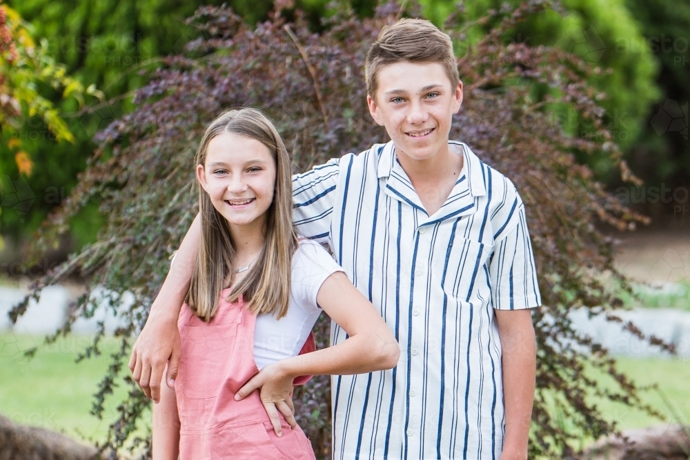 Girl standing with hand on hip with brothers arm over her shoulder smiling - Australian Stock Image