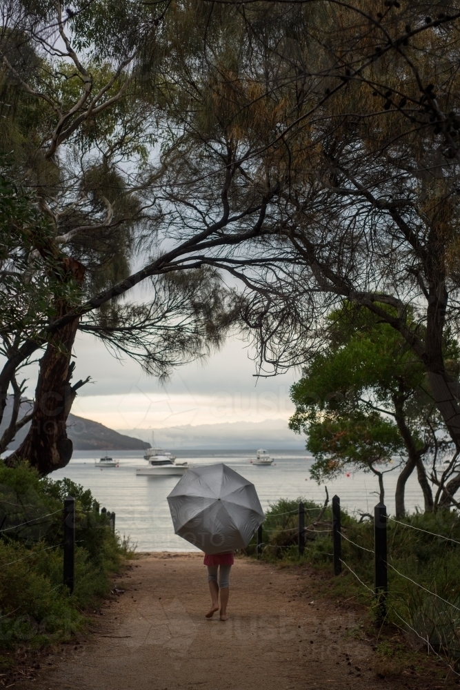 Girl walking to ocean under an umbrella surrounded by trees - Australian Stock Image