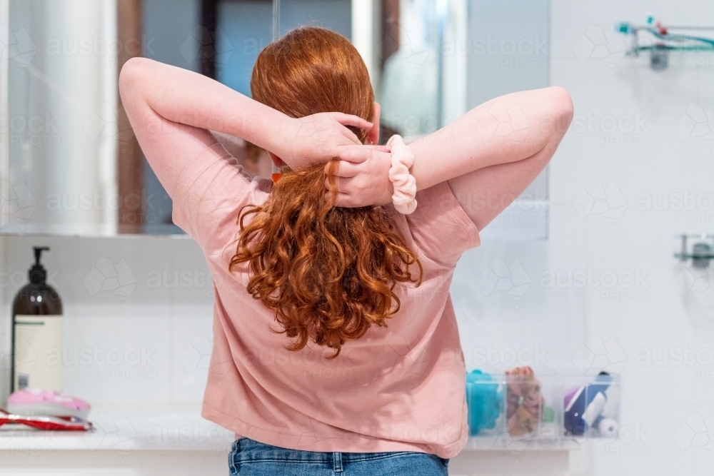 girl with fair skin and red hair in bathroom tying hair back with scrunchy - Australian Stock Image