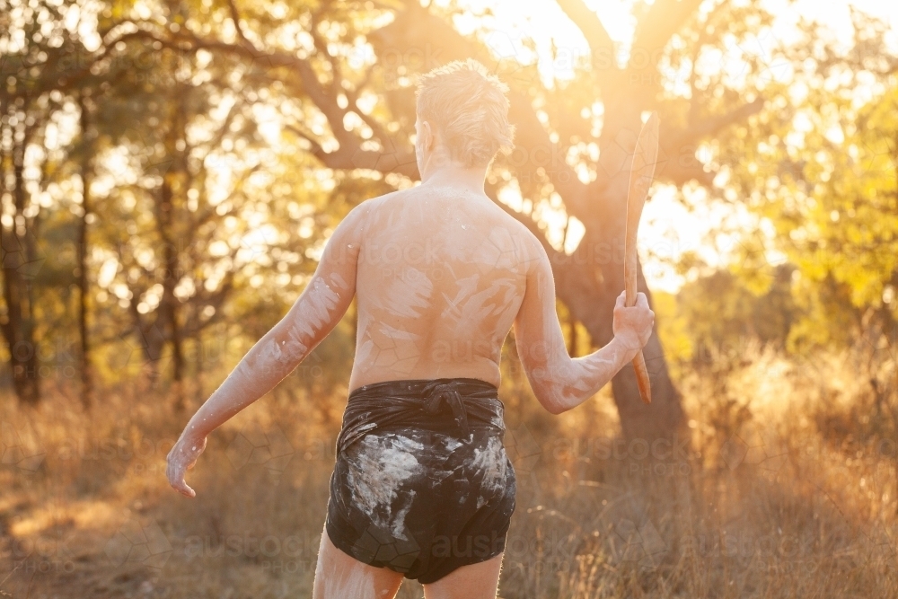 Golden Australian sun behind young First Nations man holding hunting stick in bushland - Australian Stock Image