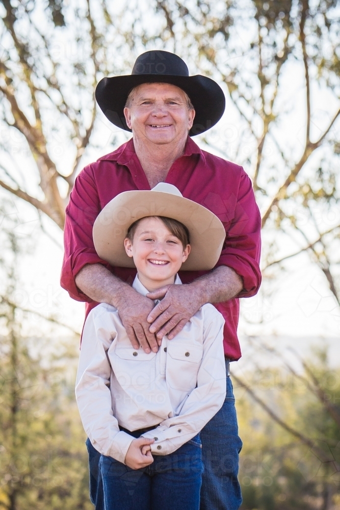 Grandfather standing with arms around grandson smiling on farm in drought - Australian Stock Image